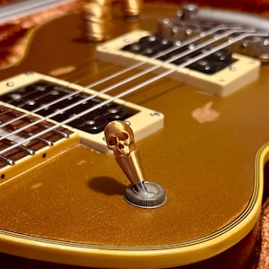 Może przedstawiać: Zbliżenie złotej gitary elektrycznej z gałką w kształcie czaszki. Gitara ma złote wykończenie z kremowymi przetwornikami i srebrnym mostkiem. Widoczne są struny, a gitara znajduje się w futerale.