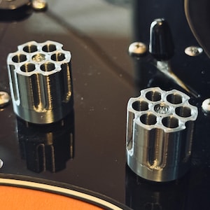 Peut inclure: Deux boutons de guitare argentés, chacun avec six trous, ressemblant à un barillet de revolver. Ils sont posés sur une surface noire brillante, avec des vis et un bouton de commande noir.