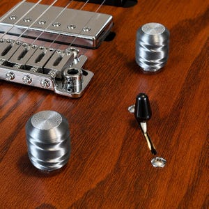 May include: A brown wooden electric guitar body featuring two cylindrical silver metal control knobs with a spiral texture, a black plastic pickup selector switch, and a chrome bridge assembly with six individual saddles and a humbucker pickup.
