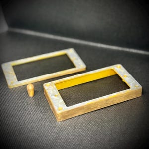 May include: Two rectangular, brass-colored frames with a yellow interior and small screw holes. A small, cylindrical brass-colored piece sits between the frames. The items are on a dark gray surface.