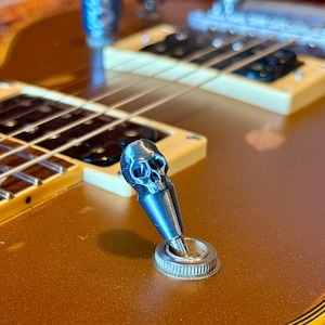May include: A silver skull-shaped guitar switch tip is mounted on a gold-coloured electric guitar. The skull features eye sockets, a nose cavity, and teeth. The guitar's strings and fretboard are visible in the background.