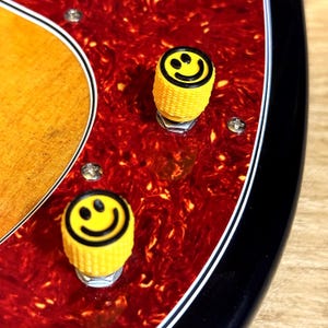 May include: Close-up of a guitar with a red and brown patterned surface. Two yellow knobs with black smiley faces are visible. The guitar has a black edge and a wooden section.