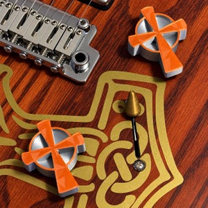 May include: A wooden electric guitar body featuring a gold-colored outline of the state of Texas with an intricate knotwork pattern inside. Two orange cross-shaped control knobs are attached, along with a metallic bridge and a small pointed switch.