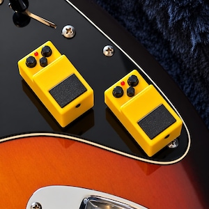 May include: Two bright yellow guitar effect pedals with black foot switches and three black control knobs each, featuring a small red indicator light. The pedals are positioned on the glossy black pickguard of a sunburst electric guitar.