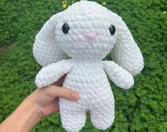 May include: A handmade white crochet bunny with long ears, black eyes, and a pink nose. The plush toy is soft and fluffy, perfect for children or as a decorative item. The bunny is held against a green background.
