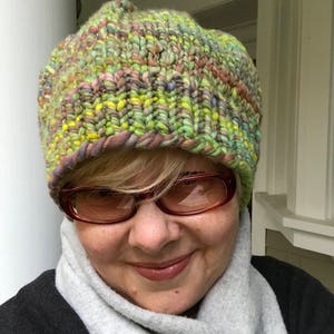 May include: A close-up of a person wearing a green, yellow, and purple knitted hat with a white scarf. The person is wearing brown-tinted glasses.