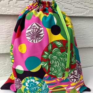 May include: A drawstring bag with a vibrant, colorful pattern. The fabric features large, overlapping circles in pink, yellow, teal, green, and black, with floral and abstract designs. A green ribbon with white polka dots and a small flower accent the top.