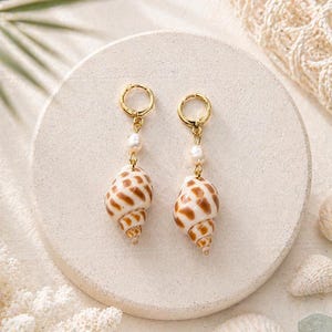 May include: A pair of gold hoop earrings with a pearl accent and a dangling seashell. The seashells are white and brown, with a spiral shape. The earrings are displayed on a white circular surface.