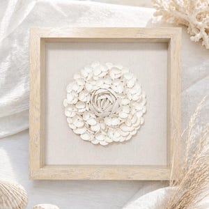 May include: A framed, square wall art piece featuring a white, floral design made of small, petal-like elements. The artwork is set against a neutral fabric background within a light-colored wooden frame. Seashells and coral are visible.