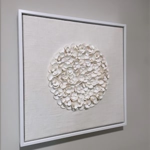 May include: Square, framed wall art featuring a circular arrangement of white, shell-like objects. The artwork is set against a light beige linen background and enclosed in a white frame. The piece is mounted on a light gray wall.