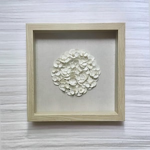 May include: A framed artwork featuring a circular arrangement of white, petal-like elements, possibly paper or fabric, with small pearl accents. The artwork is set against a neutral fabric background within a light-colored wooden frame.