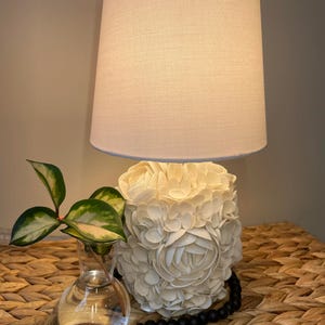 May include: A decorative table lamp with a cream-colored shade and a base adorned with cream-colored floral embellishments. A small glass vase with a green plant and a strand of black beads are next to the lamp. The lamp is on a woven surface.