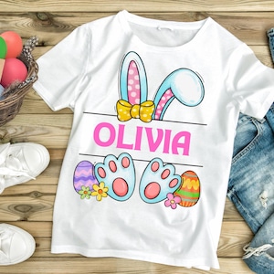 May include: White t-shirt with a cartoon bunny rabbit design. The bunny has blue ears with pink polka dots, a yellow bow, and blue paw prints. The shirt also has the name "Olivia" printed in pink letters.