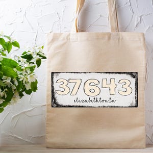Könnte beinhalten: Beige Canvas-Tragetasche mit einem schwarz-weißen rechteckigen Design. Das Design zeigt die Zahlen "37643" und den Text "elizabethton, tn" in handschriftlichem Stil. Links steht eine Vase mit weißen Blumen.