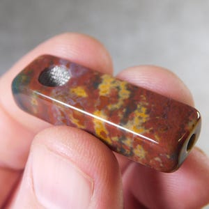 May include: A rectangular, polished stone bead with a hole drilled through it. The bead features a mix of red, yellow, and brown colors. The stone is held in a person's hand.