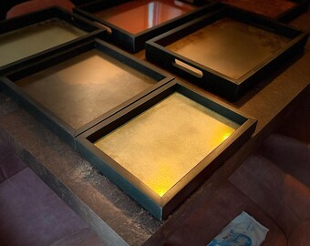 May include: Several rectangular serving trays with black frames and metallic gold, copper, and bronze interiors. The trays are arranged on a dark brown table. Each tray has a handle. The trays are in various shades of gold and brown.