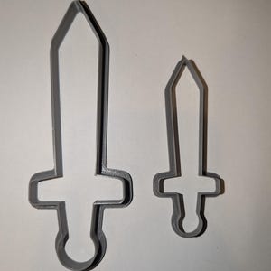 May include: Two gray plastic sword-shaped cookie cutters. One is larger than the other. The cutters have a handle and a pointed blade. The background is a plain white surface.