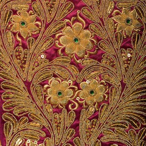 Può includere: Primo piano di un tessuto con ricami dorati intricati su uno sfondo magenta intenso. Il design presenta motivi floreali con accenti verdi e piccole paillettes, creando un aspetto lussuoso e ornato. Il ricamo è dettagliato e ricco.