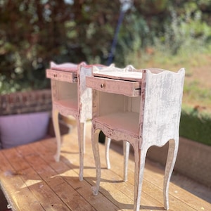 May include: A pair of matching bedside tables featuring a distressed white paint finish over wood. Each table has a single pull-out drawer with a small round knob, an open storage shelf below, and four slender, curved legs with carved detailing.