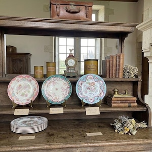 May include: A wooden hutch display featuring three decorative ceramic plates on stands, a stack of plates, and various items including a brown leather suitcase, metal canisters, a clock, books, and dried flowers. Small paper labels identify the items.