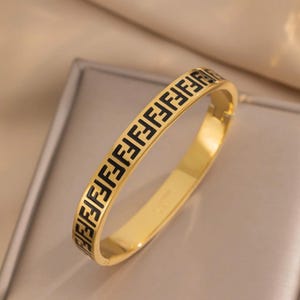 Puede incluir: Un brazalete dorado con un patrón negro repetido. El brazalete tiene un acabado liso y pulido y un diseño simple y elegante. El patrón negro está espaciado uniformemente alrededor del brazalete.
