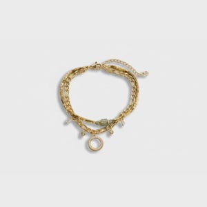 Puede incluir: Juego de pulseras doradas en capas. Las pulseras presentan varios estilos de cadena, pequeños dijes y un colgante circular con un borde texturizado. Una cuenta verde añade un toque de color. La joyería se muestra sobre un fondo blanco.
