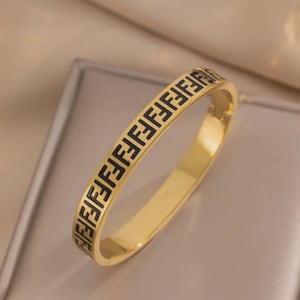 May include: A gold-toned metal bangle bracelet featuring a repeating black geometric pattern along the outer surface. The design consists of a series of interconnected letter F shapes, creating a continuous, structured motif around the entire band.