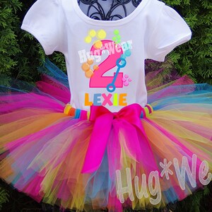 May include: A white t-shirt with a pink number two, blue and green toy, and the text "Hug*Wear" and "Lexie". The shirt is paired with a rainbow tulle tutu with a pink satin bow.