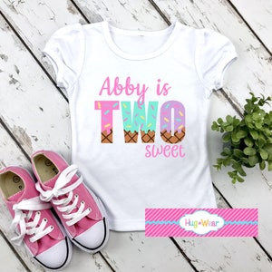 May include: White t-shirt with pink text that says "Abby is TWO sweet" with a graphic of ice cream cones with sprinkles. The shirt is on a white wooden surface with pink sneakers.