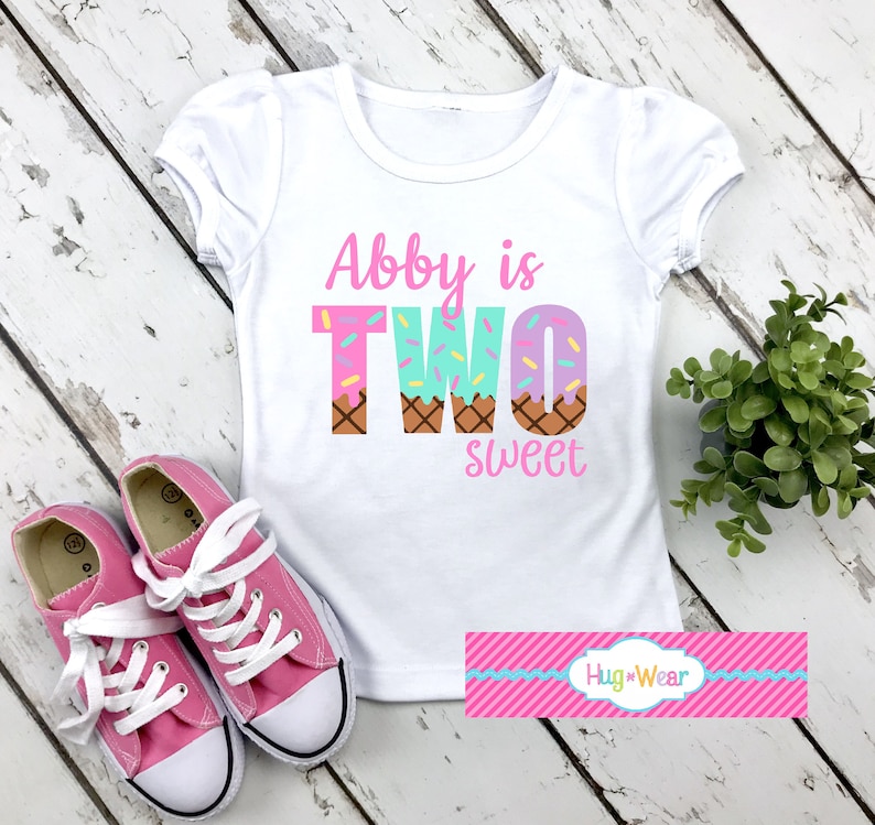 May include: White t-shirt with pink text that says "Abby is TWO sweet" with a graphic of ice cream cones with sprinkles. The shirt is on a white wooden surface with pink sneakers.