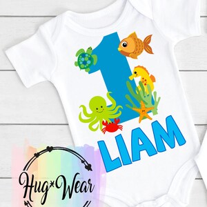 May include: White baby bodysuit with a blue number 1 and sea creature graphics. The design includes a turtle, fish, seahorse, octopus, crab, and starfish. The name "LIAM" is printed in blue. The Hug Wear logo is in the bottom left corner.