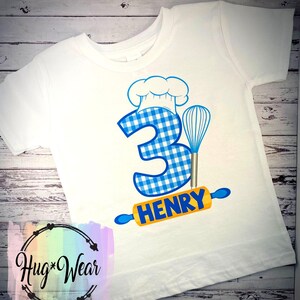 Op de afbeelding: Wit T-shirt met een blauw-wit geruit nummer 3, een koksmuts, een deegroller en een garde. De tekst "HENRY" staat op de deegroller.