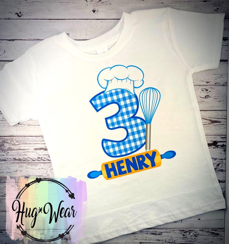 Op de afbeelding: Wit T-shirt met een blauw-wit geruit nummer 3, een koksmuts, een deegroller en een garde. De tekst "HENRY" staat op de deegroller.