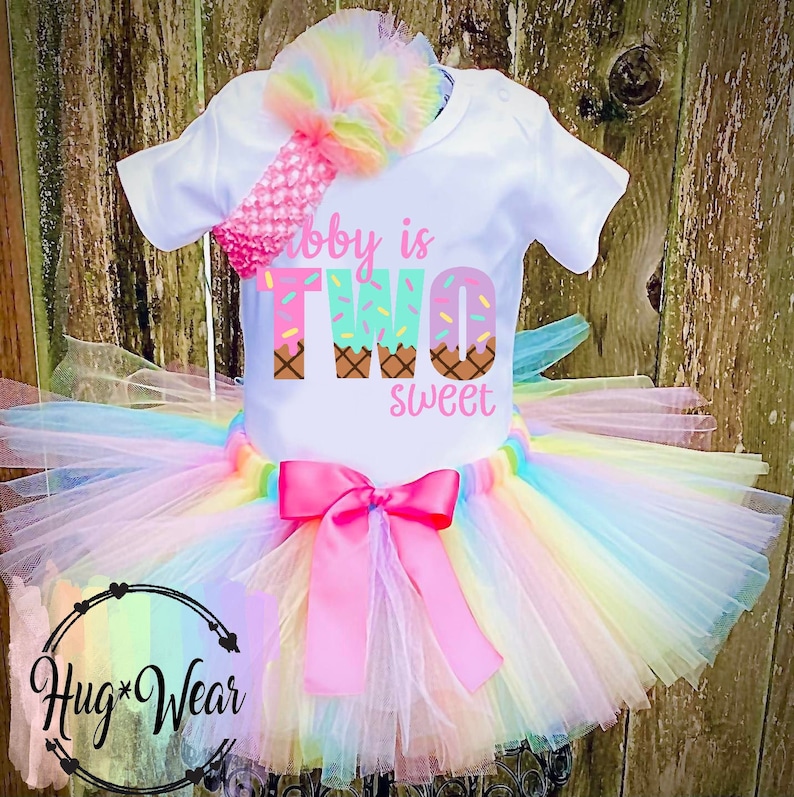 May include: A white onesie with the text "Baby is Two Sweet" in pink, green, and brown. The number two is decorated with ice cream cones. The onesie is paired with a rainbow tulle tutu and a pink headband.