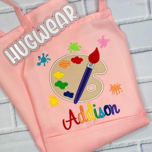 Pu&ograve; includere: Grembiule da artista rosa per bambini con un design di tavolozza e pennello. Il grembiule ha una tasca e il testo "Hugwear" e "Addison" sono ricamati sul davanti.