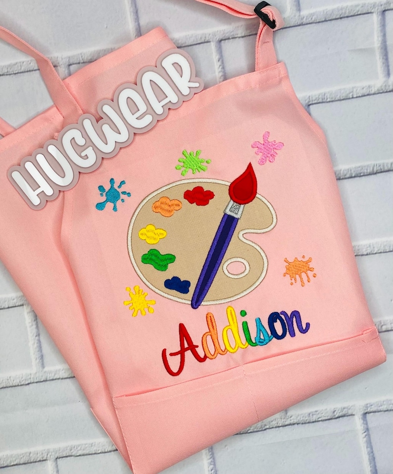Pu&ograve; includere: Grembiule da artista rosa per bambini con un design di tavolozza e pennello. Il grembiule ha una tasca e il testo "Hugwear" e "Addison" sono ricamati sul davanti.