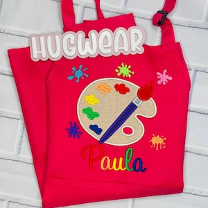 Pu&ograve; includere: Grembiule da artista rosa per bambini con un design di tavolozza e pennello. Il grembiule &egrave; ricamato con le parole "Hugwear" e "Paula".