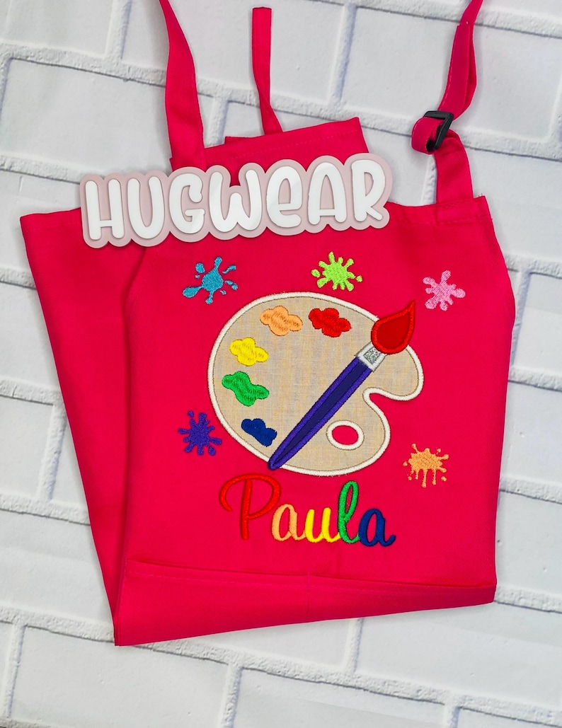 Pu&ograve; includere: Grembiule da artista rosa per bambini con un design di tavolozza e pennello. Il grembiule &egrave; ricamato con le parole "Hugwear" e "Paula".