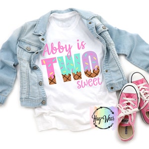 May include: White t-shirt with the text "Abby is Two sweet" in pink and blue letters. The letters are decorated with sprinkles and ice cream cone designs.