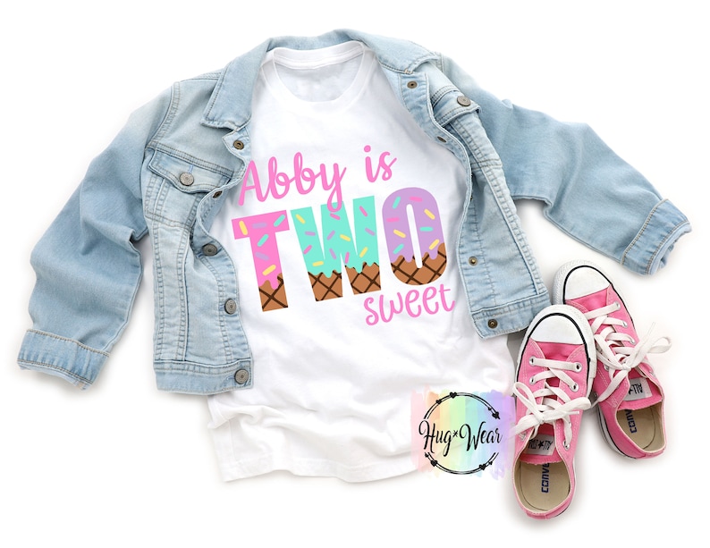 May include: White t-shirt with the text "Abby is Two sweet" in pink and blue letters. The letters are decorated with sprinkles and ice cream cone designs.