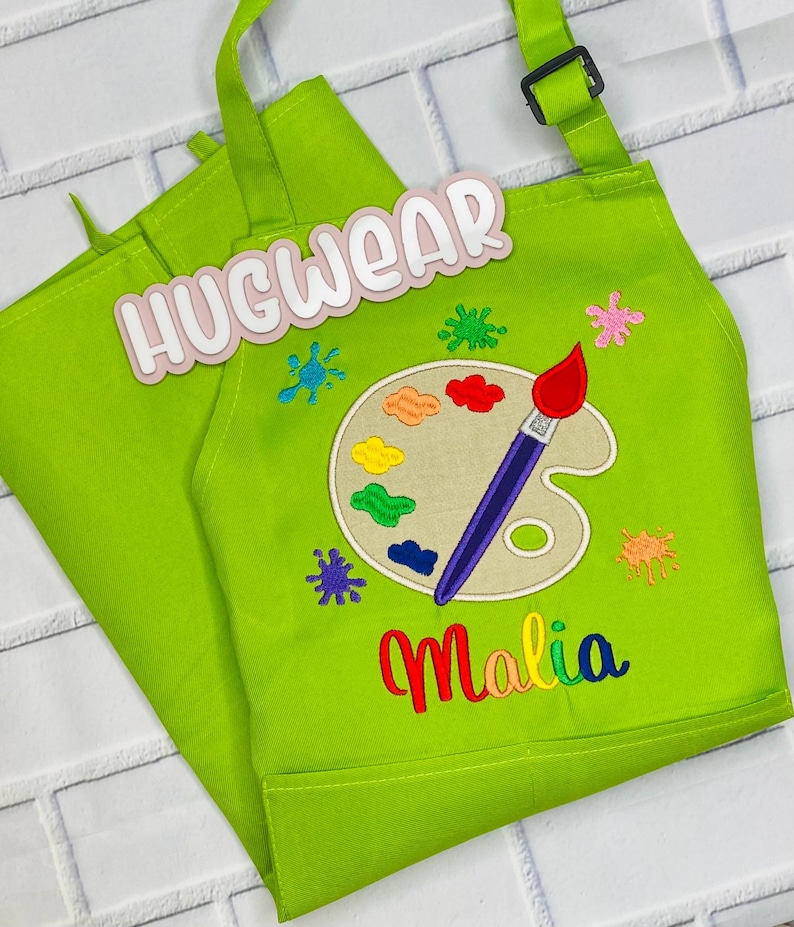 Pu&ograve; includere: Un grembiule verde per bambini con un design a forma di tavolozza e pennello. Il grembiule ha una tasca e spalline regolabili. Il testo "Hugwear" e "Malia" &egrave; ricamato sul grembiule.