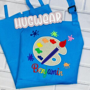 Pu&ograve; includere: Un grembiule da arte blu per bambini con un design di tavolozza e pennello. Il grembiule ha il testo "Hugwear" e "Benjamin" ricamato.