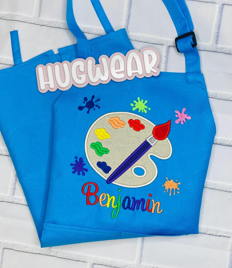Pu&ograve; includere: Un grembiule da arte blu per bambini con un design di tavolozza e pennello. Il grembiule ha il testo "Hugwear" e "Benjamin" ricamato.