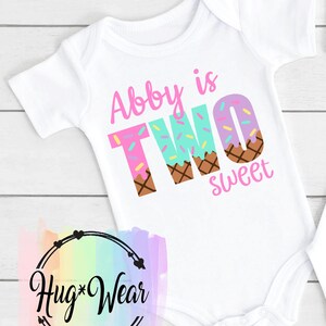 May include: White baby onesie with pink, blue, and brown ice cream cone letters spelling "Abby is TWO sweet".