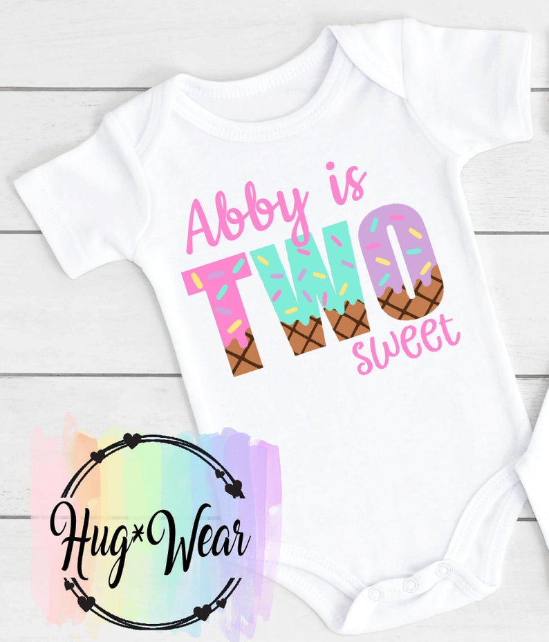 May include: White baby onesie with pink, blue, and brown ice cream cone letters spelling "Abby is TWO sweet".
