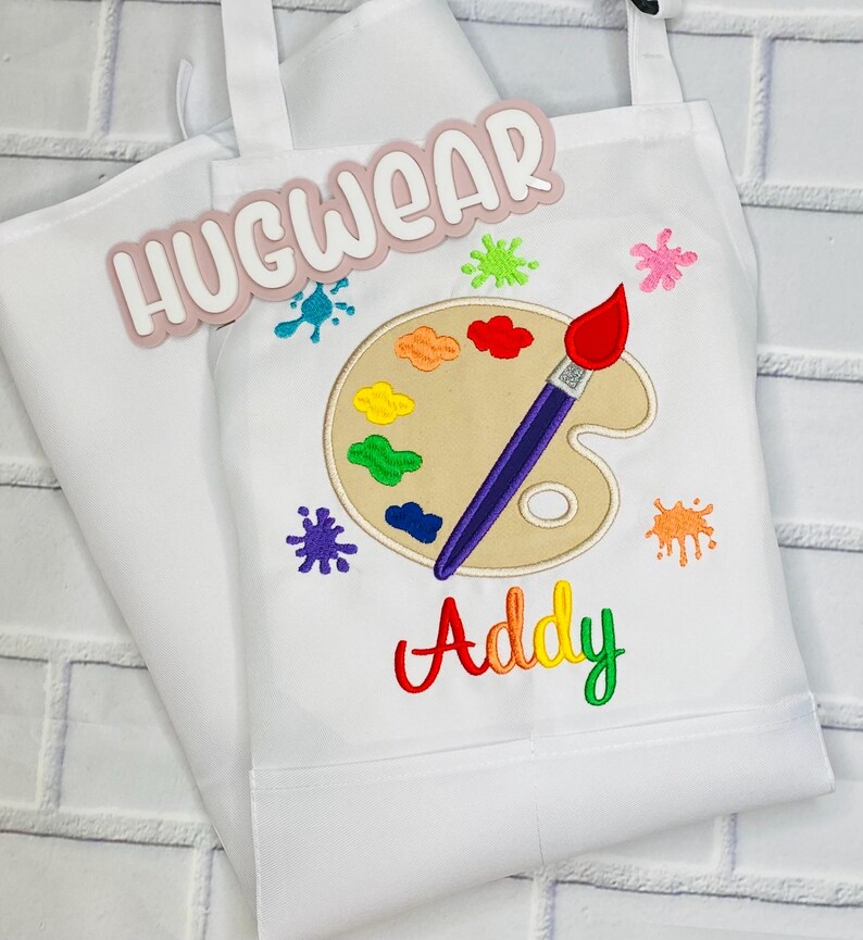 Pu&ograve; includere: Grembiule bianco con un design di tavolozza e pennello. Il testo "Hugwear" e "Addy" sono ricamati sul grembiule.