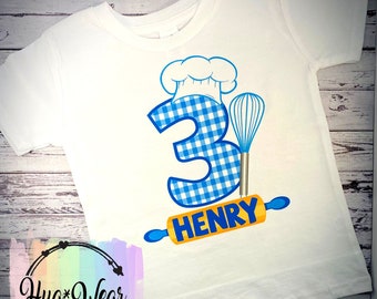 May include: White t-shirt with a blue and white checkered number 3, a blue chef's hat, a rolling pin, and a whisk. The rolling pin has the name "HENRY" on it.