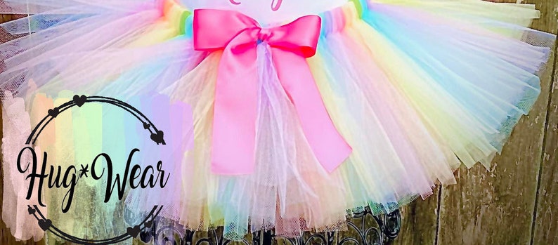 May include: A rainbow tulle tutu with a pink satin bow. The tutu is made of multiple layers of tulle and has a rainbow ombre effect. The bow is tied in the center of the tutu and is a bright pink color.