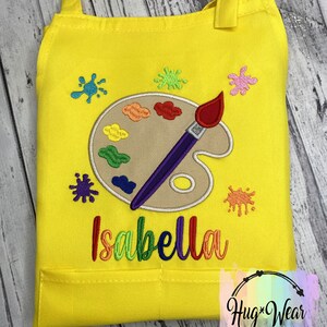 Pu&ograve; includere: Grembiule da artista giallo per bambini con un design di tavolozza e pennello. Il grembiule ha una tasca e il nome "Isabella" &egrave; ricamato sul davanti.