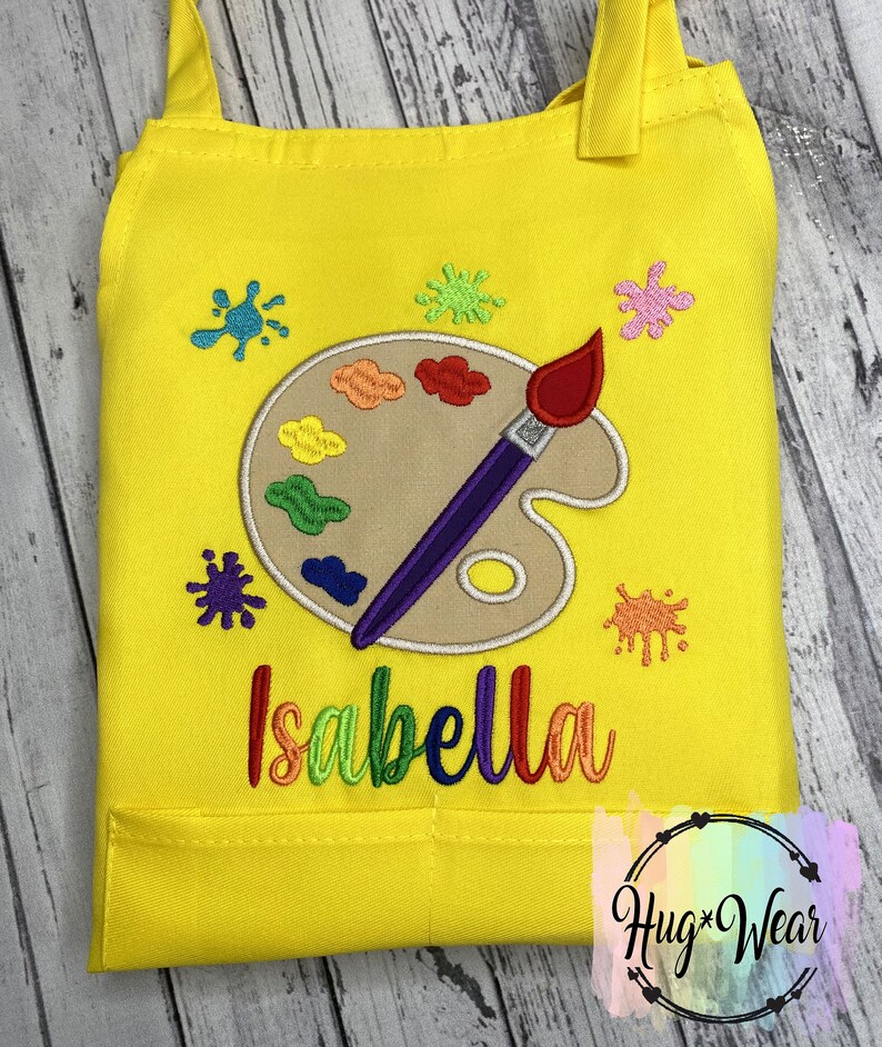 Pu&ograve; includere: Grembiule da artista giallo per bambini con un design di tavolozza e pennello. Il grembiule ha una tasca e il nome "Isabella" &egrave; ricamato sul davanti.
